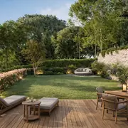 Aménagement extérieur comprenant une vaste plage en bois et un espace verdoyant. Un muret en pierre naturelle délimite le terrain arboré propice au repos.